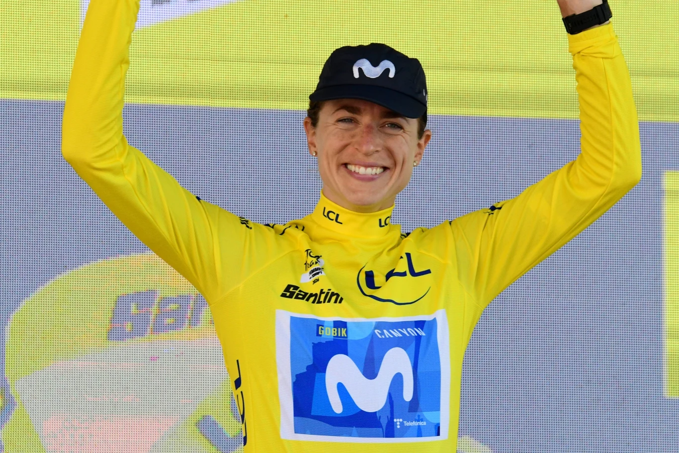 Marlen Reusser Kolarz w żółtej koszulce zwycięzcy z logo zespołu Movistar, unoszący ręce w geście triumfu na tle żółtej ścianki sponsorskiej.
