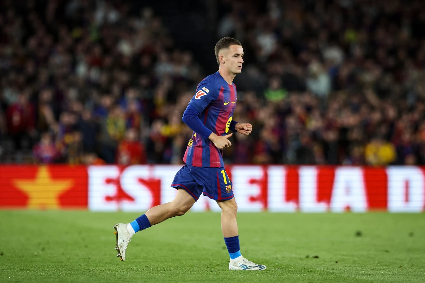 Piłkarz w koszulce FC Barcelony biegnący po murawie stadionu podczas meczu, w tle widownia i rozmyte światła reklamowe.