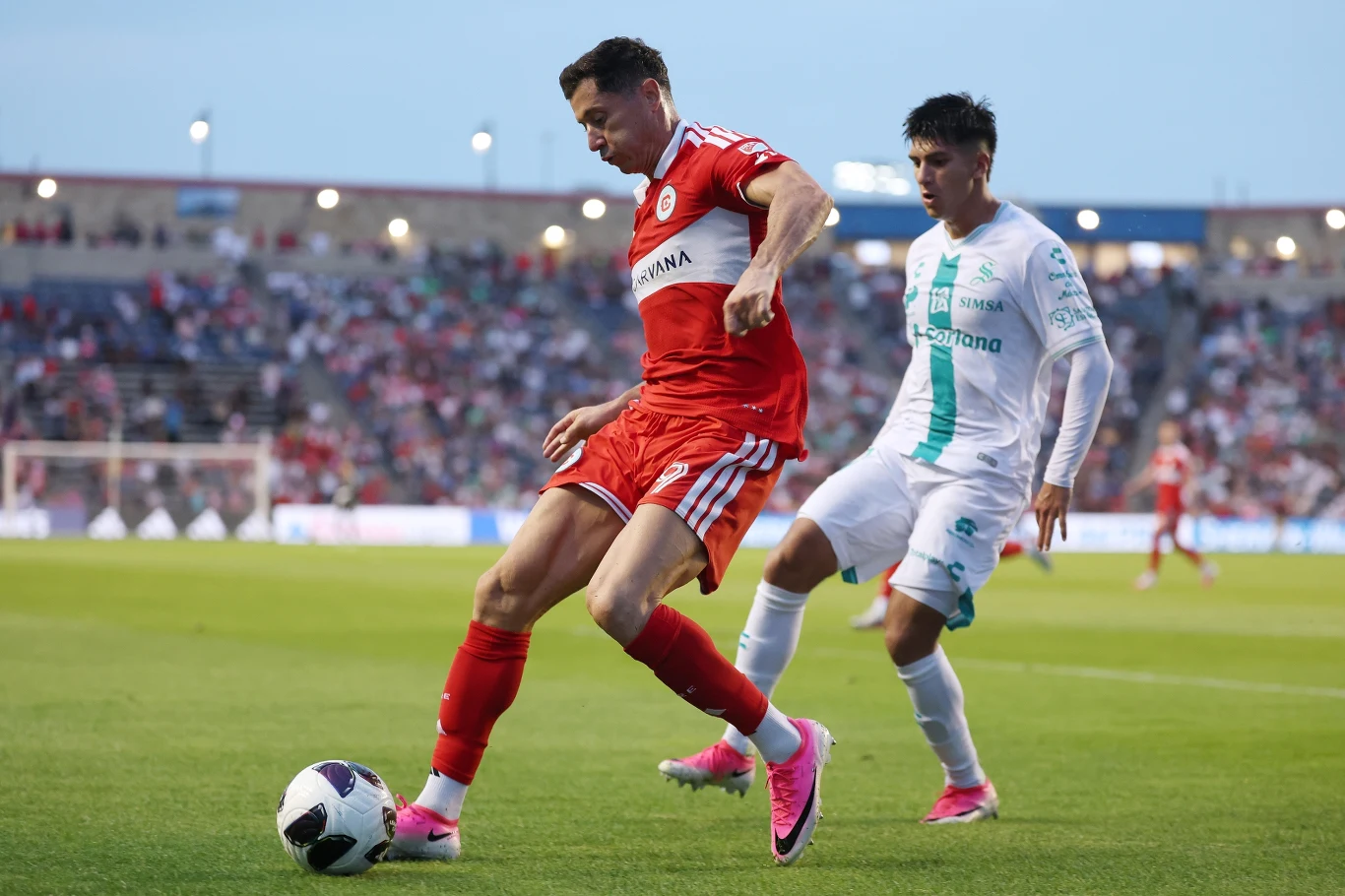 Kadr z meczu Chicago Fire - Santos Laguna, na pierwszym planie Robert Lewandowski Dwóch piłkarzy walczy o piłkę podczas meczu na stadionie; zawodnik w czerwonym stroju kontroluje futbolówkę, a rywal w białozielonym stroju próbuje go powstrzymać.