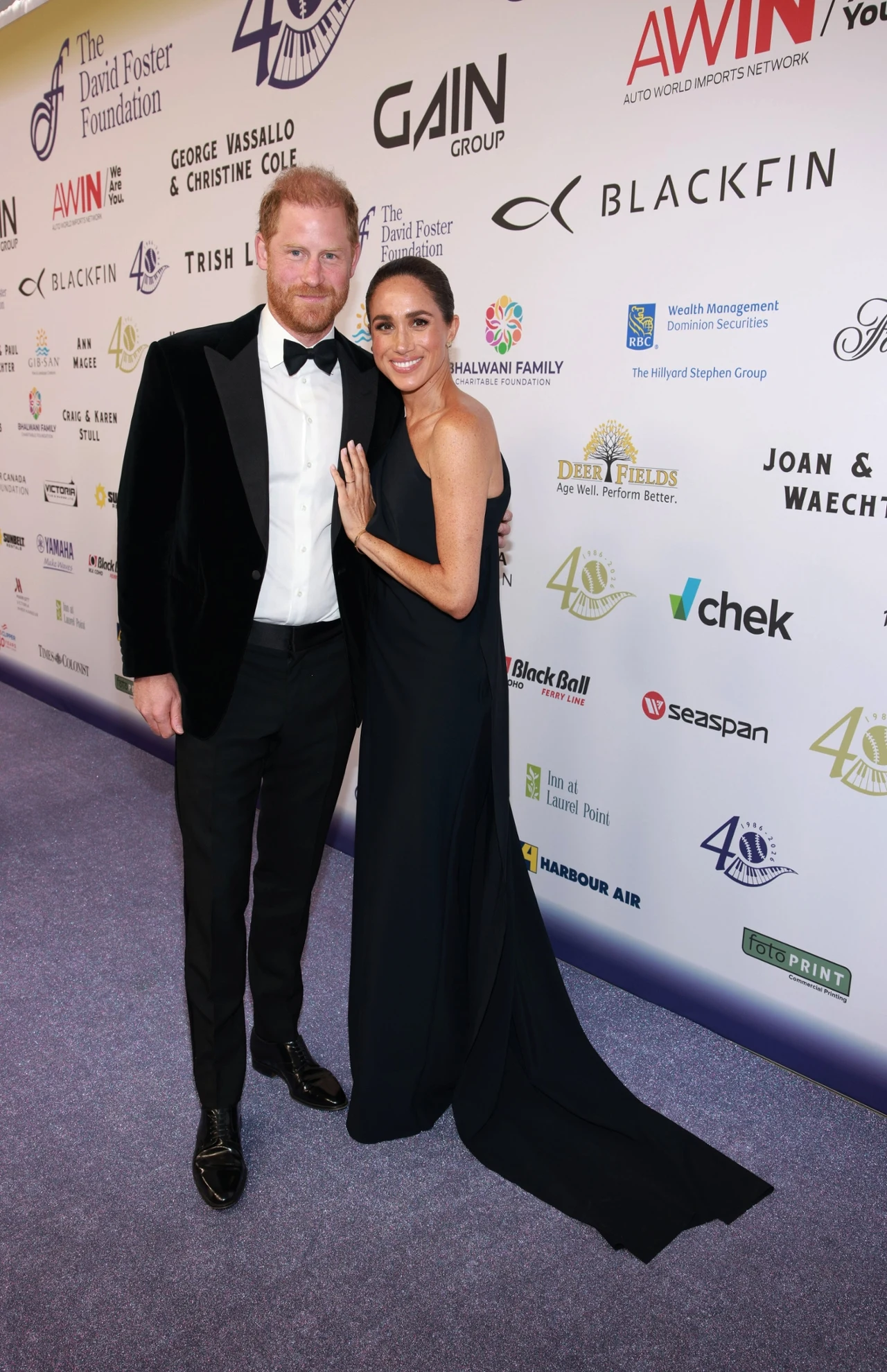 Spektakularna suknia zdecydowanie dodawała klasy Meghan Para ubrana w eleganckie wieczorowe stroje pozuje razem na tle ściany z logotypami sponsorów podczas uroczystego wydarzenia.