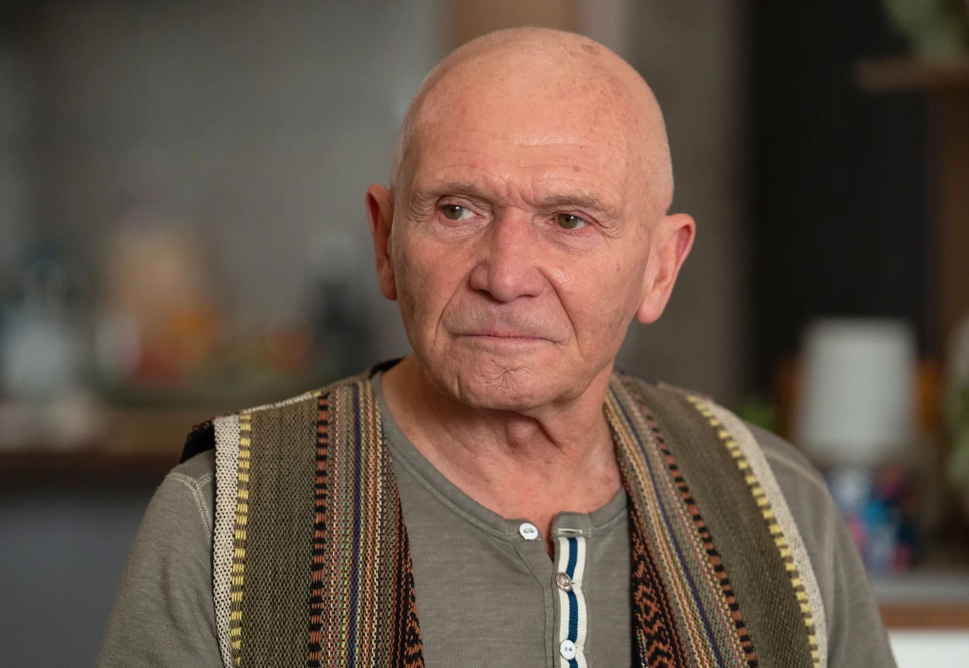 Starszy mężczyzna o krótkich, siwych włosach, ubrany w szarą koszulkę z guzikiem pod szyją oraz we wzorzysty, kolorowy sweter zarzucony na ramiona, siedzi w pomieszczeniu z rozmytym tłem.