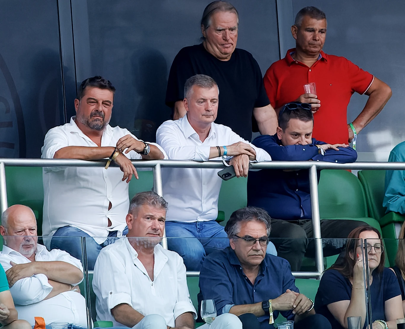 Pal Orosz i Gabor Kubatov, 19.07.2023 r. Grupa mężczyzn siedzących i stojących na trybunach stadionu, większość z nich obserwuje wydarzenie sportowe z poważnymi lub zamyślonymi minami, niektórzy opierają się o barierki.