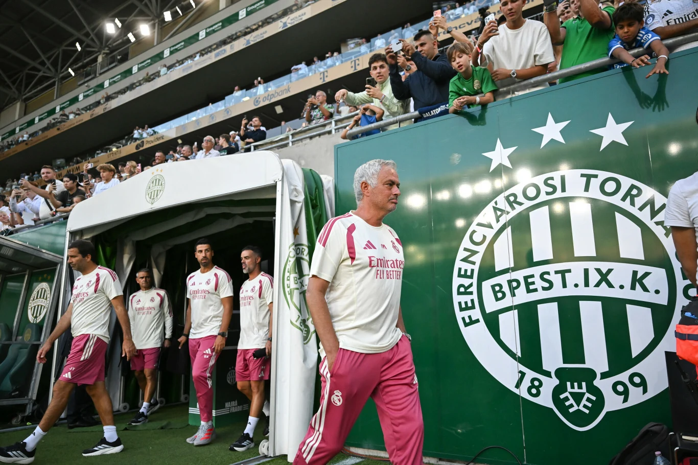 Jose Mourinho na stadionie Ferencvarosu, 08.08.2026 r. Trener drużyny piłkarskiej w jasnoróżowym stroju wychodzi z tunelu stadionu, a za nim podążają zawodnicy; obok widoczny duży herb klubu Ferencvárosi TC oraz tłum kibiców obserwujących sytuację zza ogrodzenia.