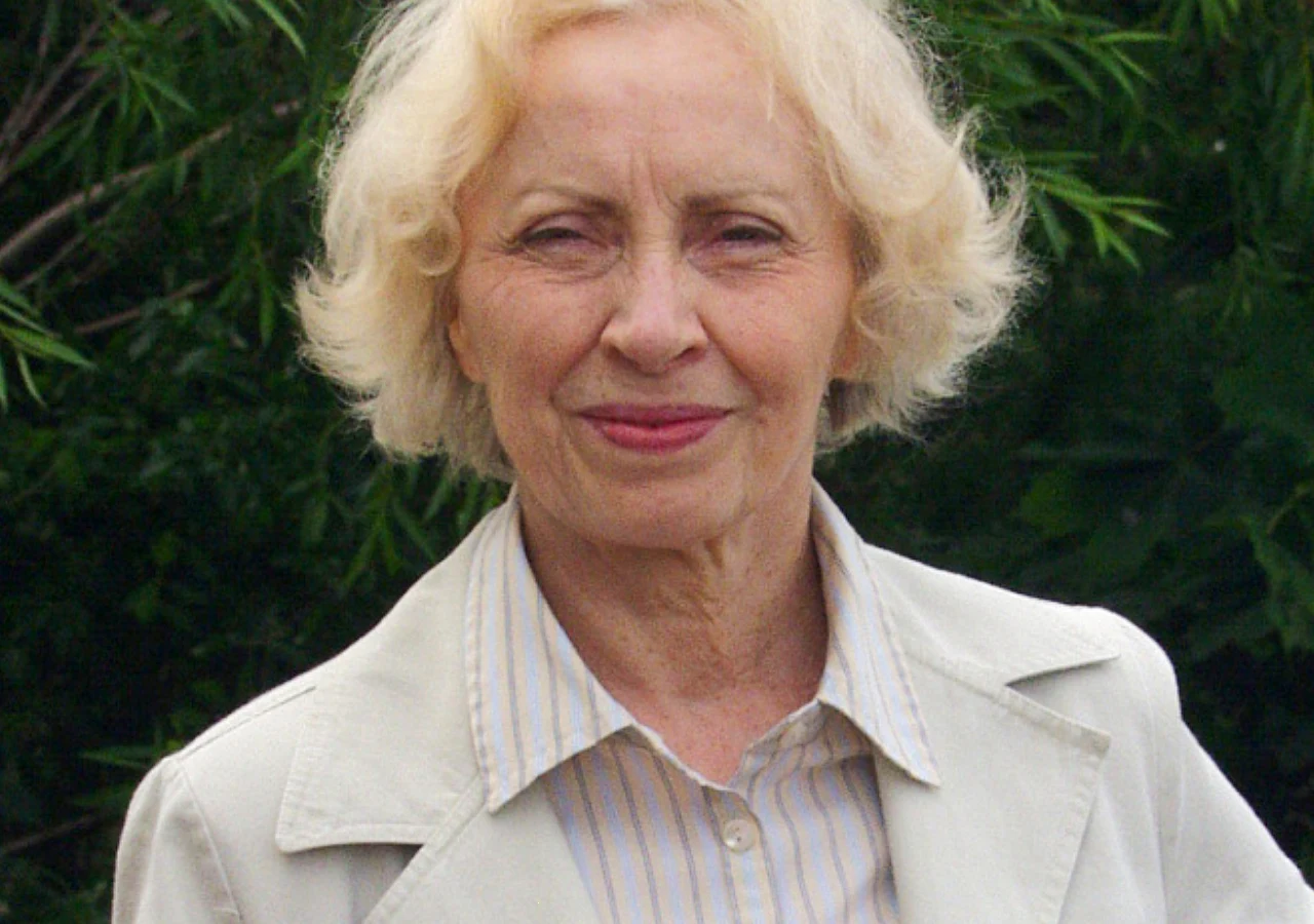 Anna Polony (2002 rok) Starsza kobieta o jasnych, kręconych włosach, ubrana w jasną marynarkę i koszulę w paski, stoi na tle zieleni.