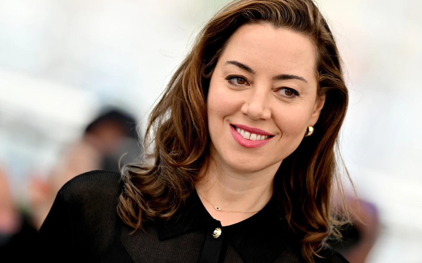 Aubrey Plaza Aubrey Plaza w czarnej bluzce, złotych kolczykach i naszyjniku, patrzy w bok na jasnym tle.