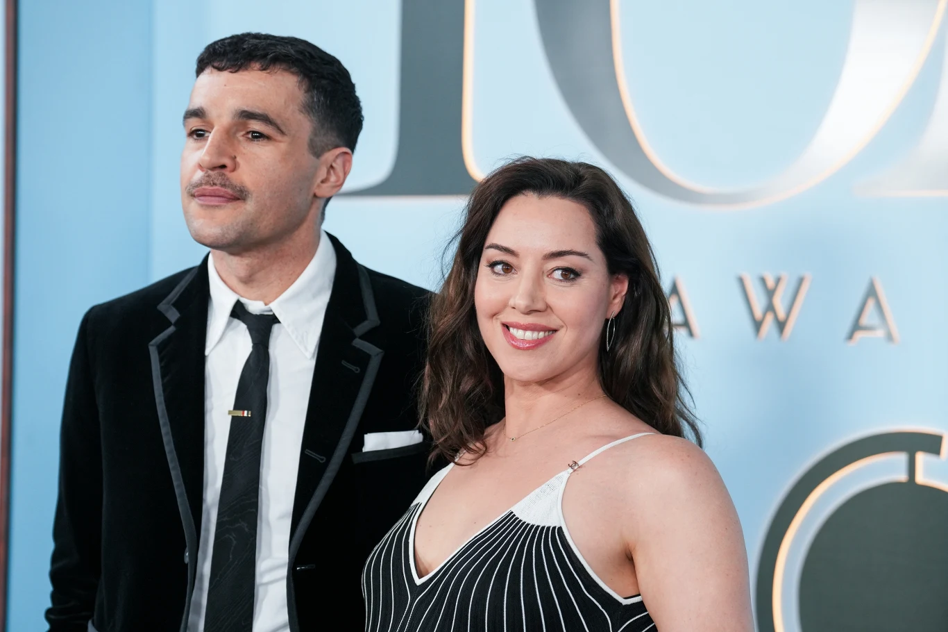 Christopher Abbott i Aubrey Plaza Para ubrana w eleganckie stroje wieczorowe stoi razem na tle niebieskiej ścianki z dużymi literami, kobieta uśmiecha się do aparatu, a mężczyzna patrzy lekko w bok.