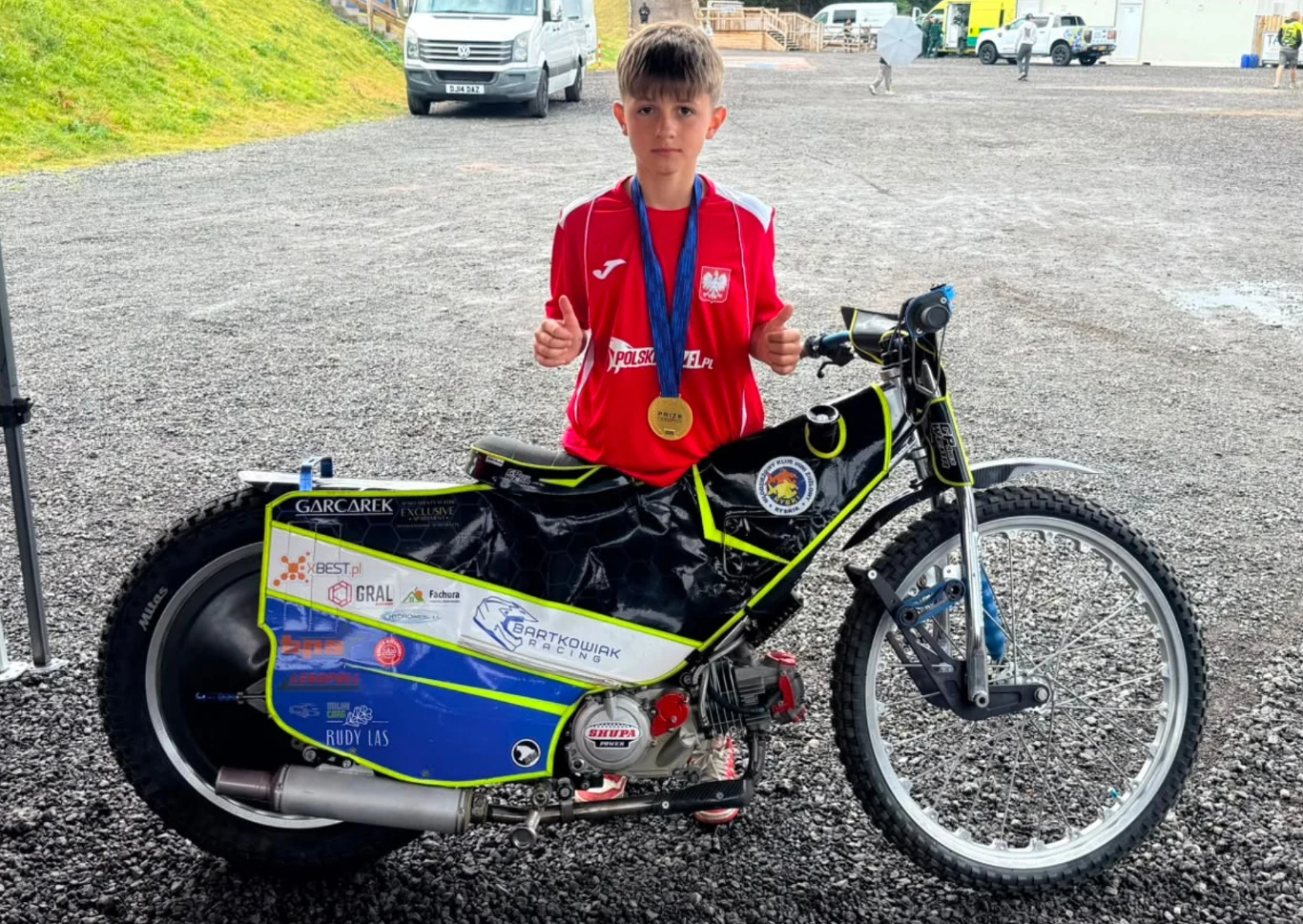 Chłopiec w czerwonej koszulce sportowej z medalem na szyi stoi za motocyklem żużlowym, trzymając kciuki uniesione do góry.