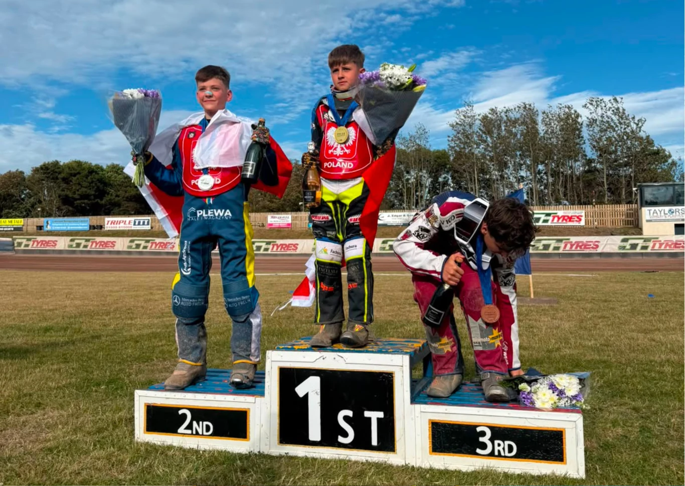 Trzech młodych żużlowców na podium; zwycięzca w środku w stroju z flagą Polski trzyma medal, bukiet kwiatów i butelkę szampana, zdobywcy drugiego i trzeciego miejsca stoją po bokach również z medalami i kwiatami.