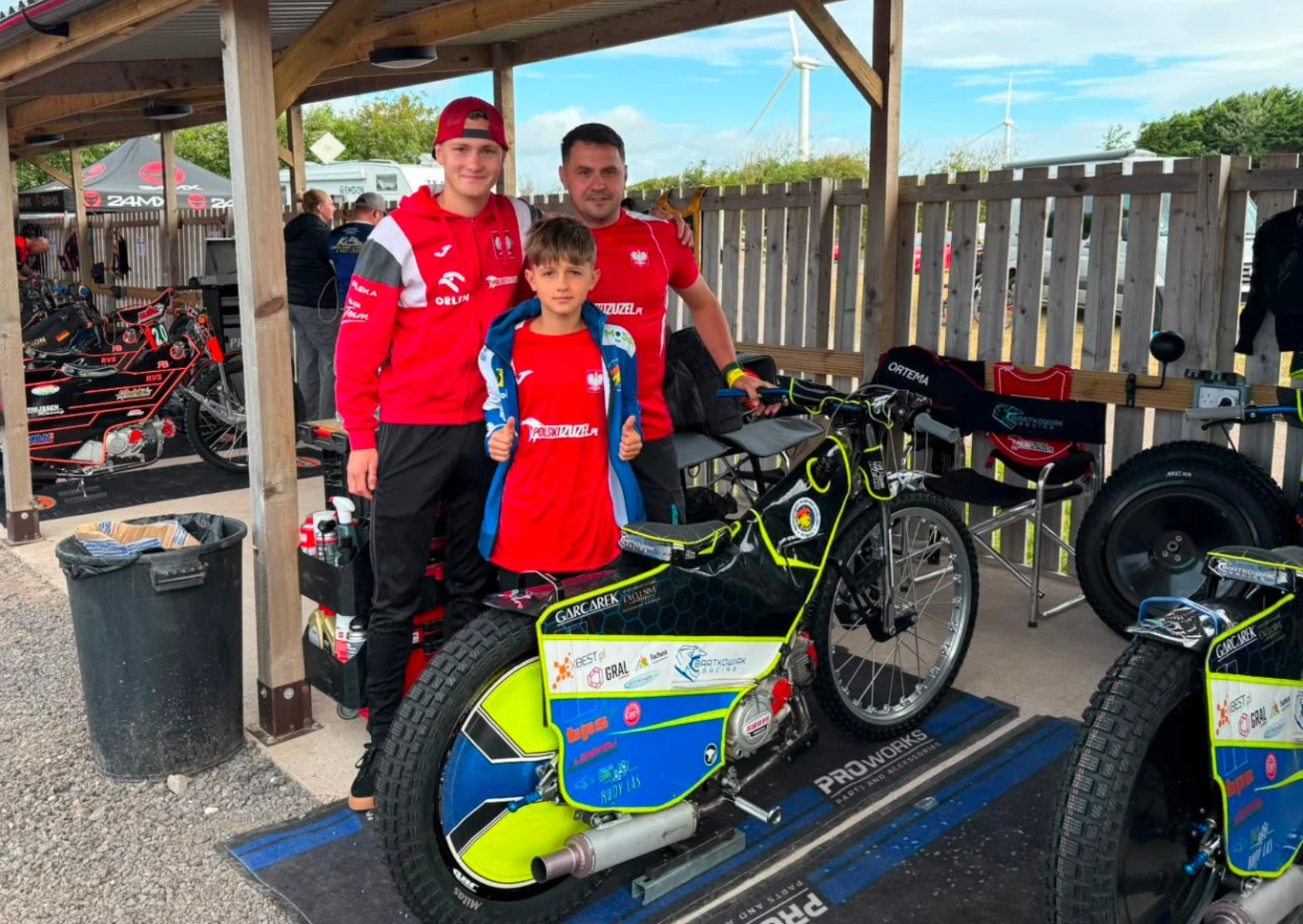 Trzech chłopców w czerwonych koszulkach drużynowych pozuje obok motocykla żużlowego w strefie serwisowej toru wyścigowego.