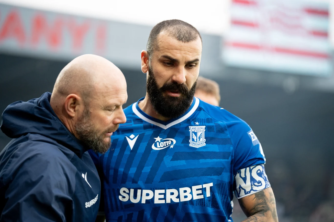 Mikael Ishak Piłkarz w niebieskim stroju z opaską kapitańską stoi obok trenera, obaj skoncentrowani na rozmowie podczas wydarzenia sportowego.