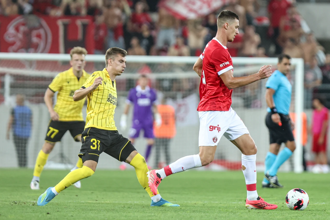 Hapoel Tel Awiw - GKS Katowice w III rundzie eliminacji Ligi Konferencji Zawodnik w czerwono-białym stroju prowadzi piłkę, naciskany przez przeciwnika w żółto-czarnym stroju podczas meczu piłkarskiego, otoczeni innymi piłkarzami i sędzią na tle trybun wypełnionych kibicami.