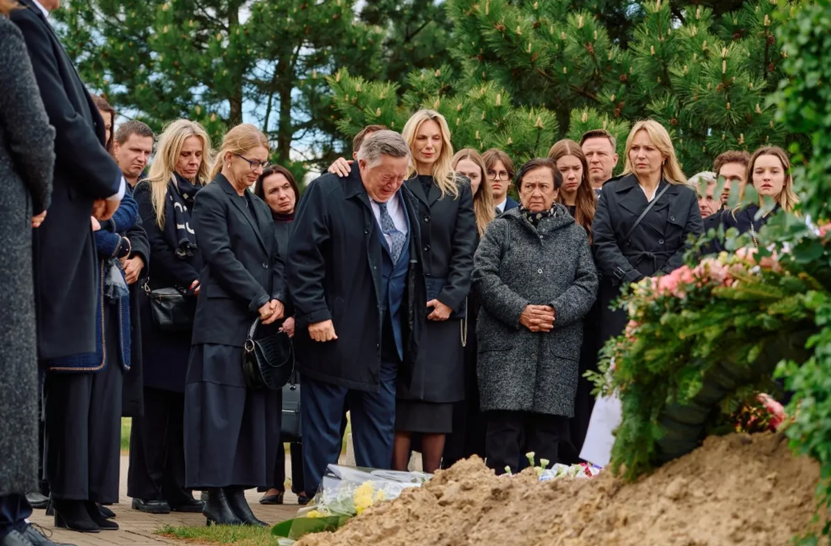 Grupa ludzi ubrana na czarno stoi w skupieniu wokół świeżo usypanego grobu, wyrażając smutek i żałobę, obok widoczne są kwiaty i wiązanki pogrzebowe.