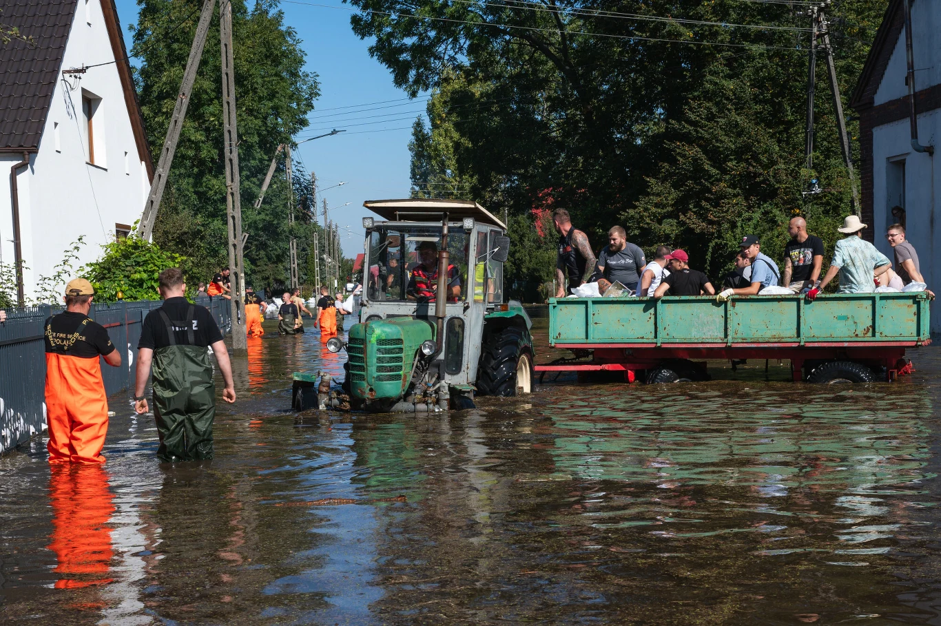 Fala powodziowa w Ścinawie, 21.09.2024 Traktor z przyczepą przewożący ludzi przez zalaną ulicę w Ścinawie, 21.09.2024, otoczony budynkami i osobami w wodoodpornych ubraniach.