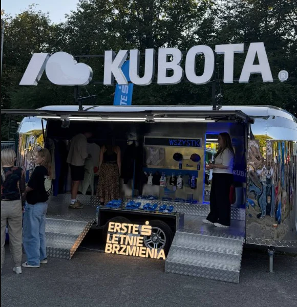 Stoisko z charakterystycznym logo marki Kubota, mobilny punkt obsługujący uczestników wydarzenia z gadżetami i produktami, przy którym gromadzą się zainteresowane osoby.