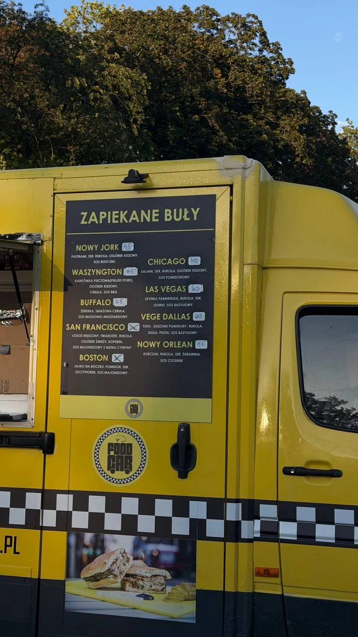 Menu z różnymi rodzajami zapiekanych buł widoczne na boku żółtego food trucka, ozdobionego czarno-białą szachownicą i ilustracją przekrojonej bułki z dodatkami.