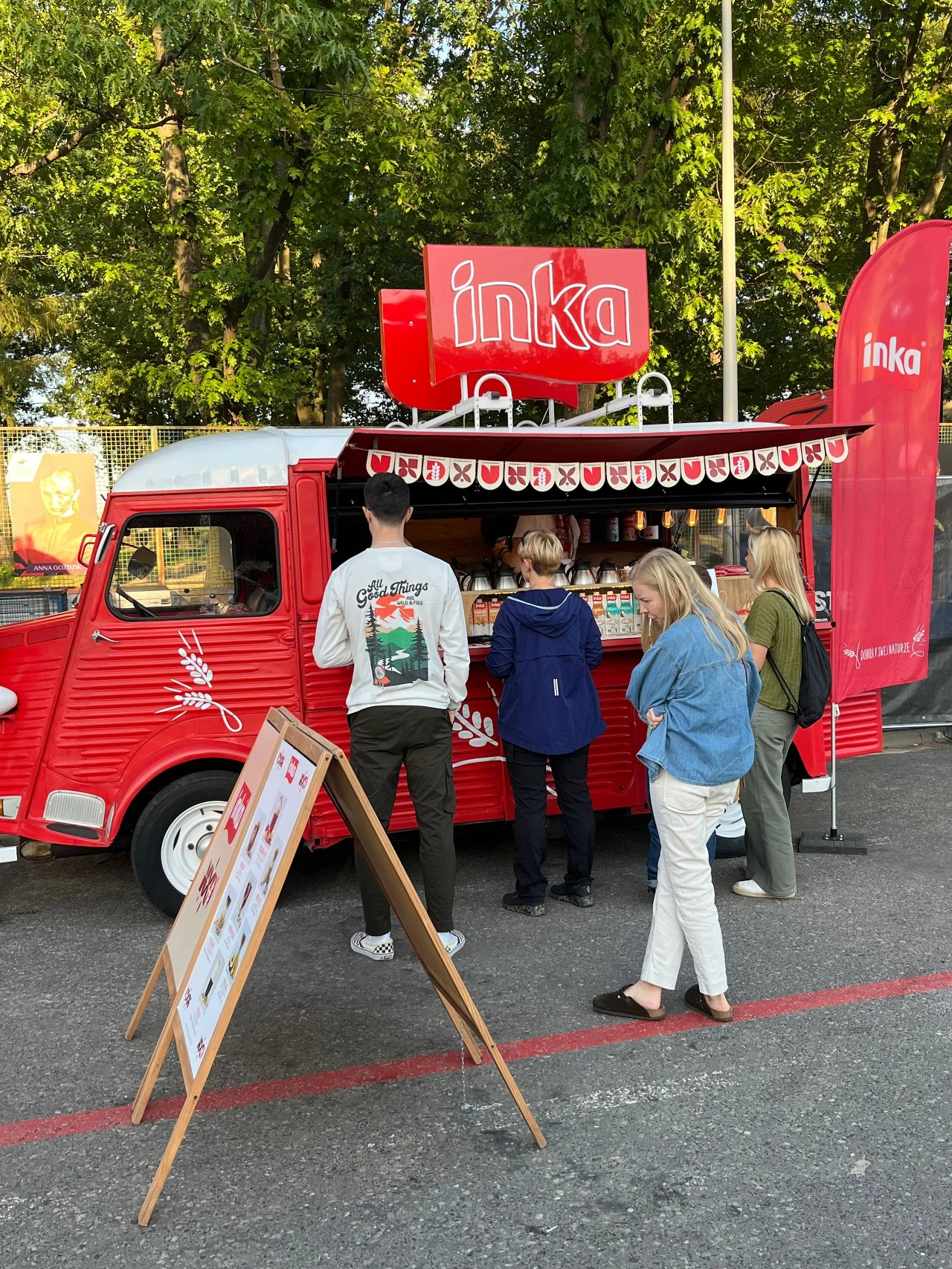 Czerwony food truck marki Inka serwujący napoje i przekąski, przy którym stoi grupa osób oczekujących na zamówienie, na pierwszym planie widoczna tablica z menu.