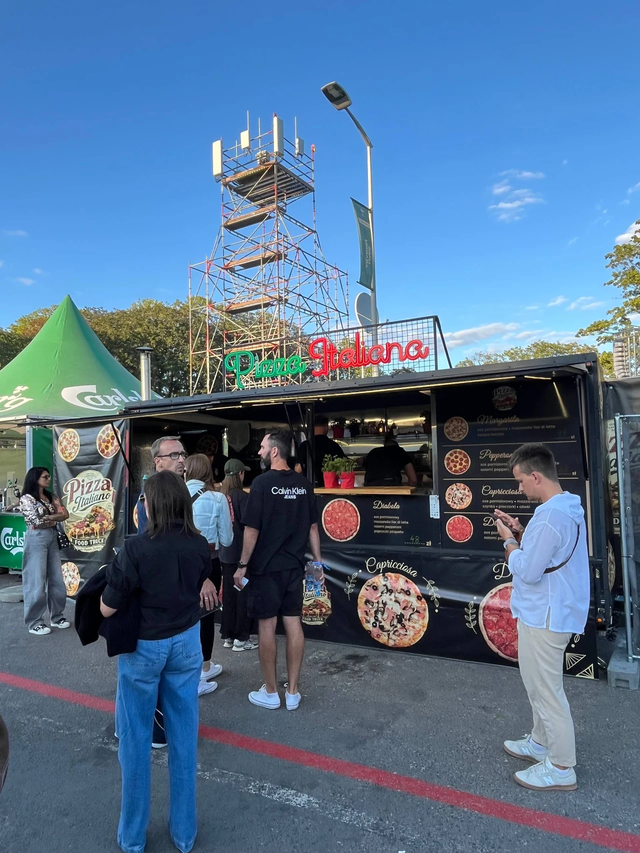 Grupa osób czeka przy czarnym food trucku serwującym pizzę, z jasnymi zdjęciami różnych rodzajów pizzy na froncie stoiska; w tle widoczna metalowa konstrukcja wieży oraz zielony namiot firmowy.