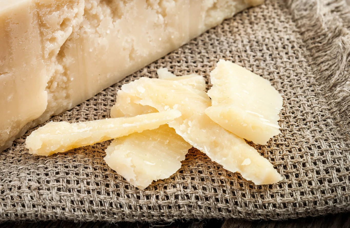 Parmezan to smaczny, słony ser bogaty w białko i minerały Kawałki twardego, złocistego parmezanu na szarej tkaninie, obok większego bloku sera o nieregularnej strukturze.