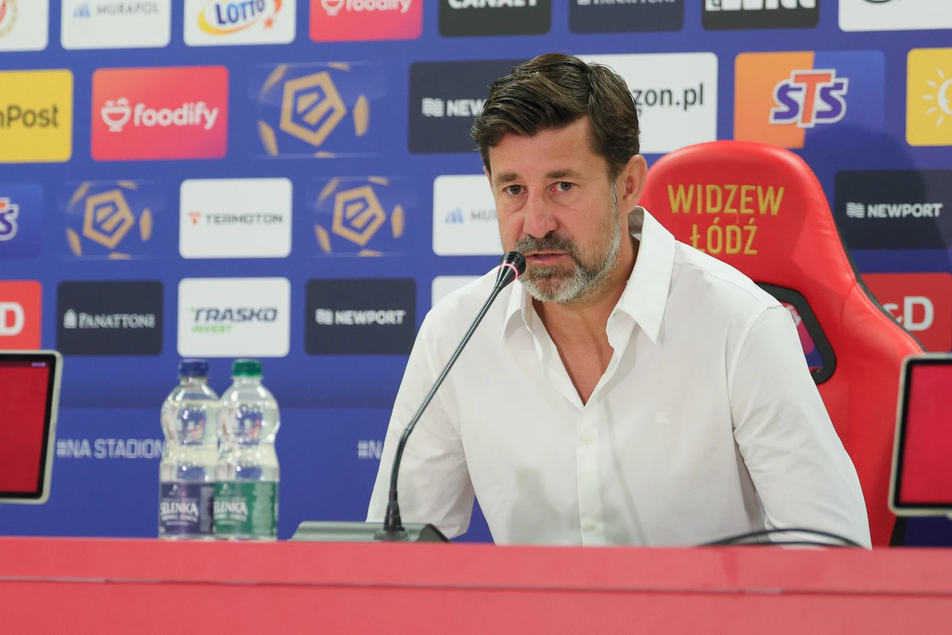 Łukasz Masłowski Mężczyzna w białej koszuli siedzi przy stole konferencyjnym z mikrofonem, na tle ściany wypełnionej logotypami sponsorów oraz nazwą klubu Widzew Łódź, obok stoją dwie butelki wody.