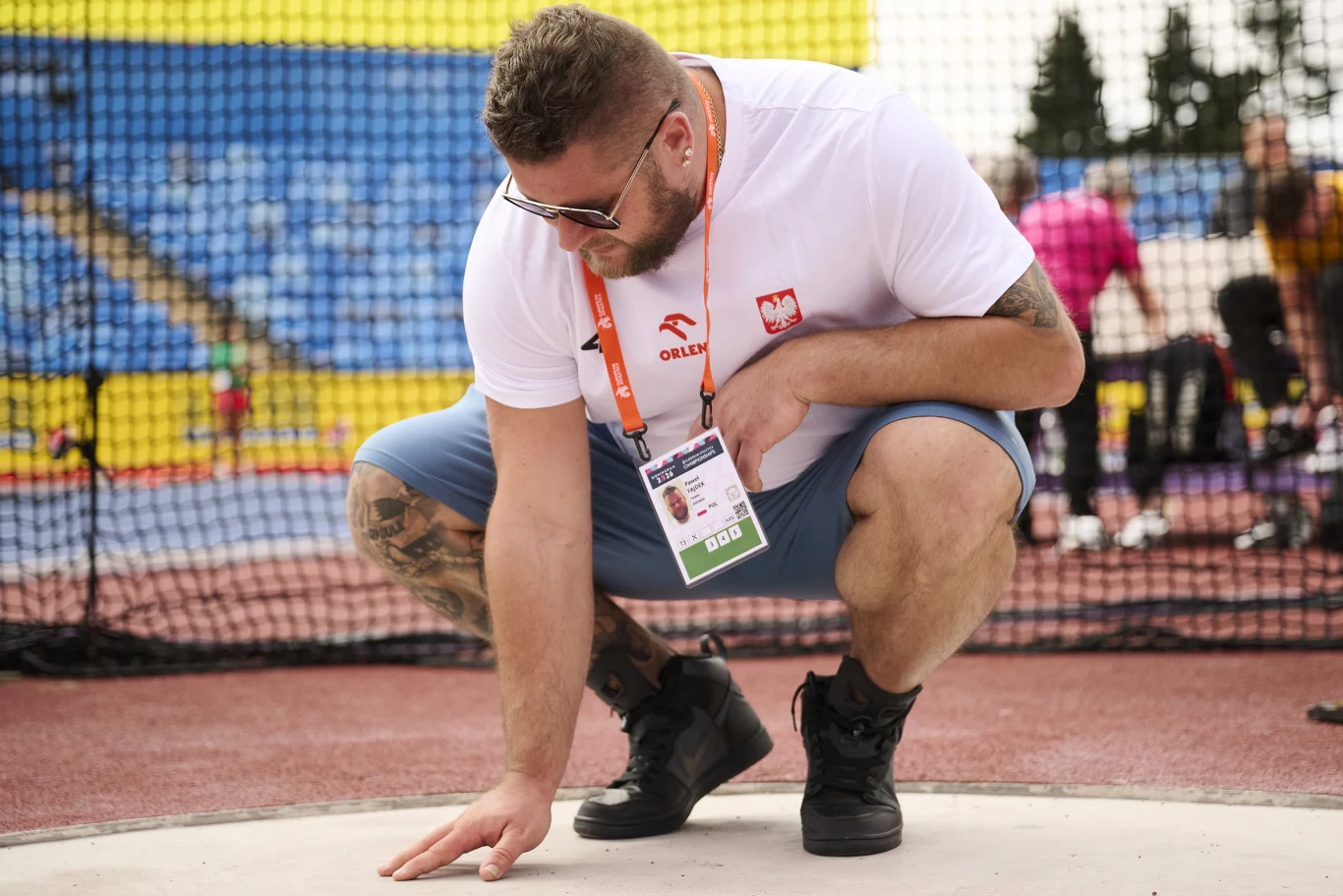 Paweł Fajdek sprawdza koło w Birmingham Mężczyzna w białej koszulce z polskim godłem i identyfikatorem pochyla się, dotykając ręką powierzchni koła na stadionie lekkoatletycznym.