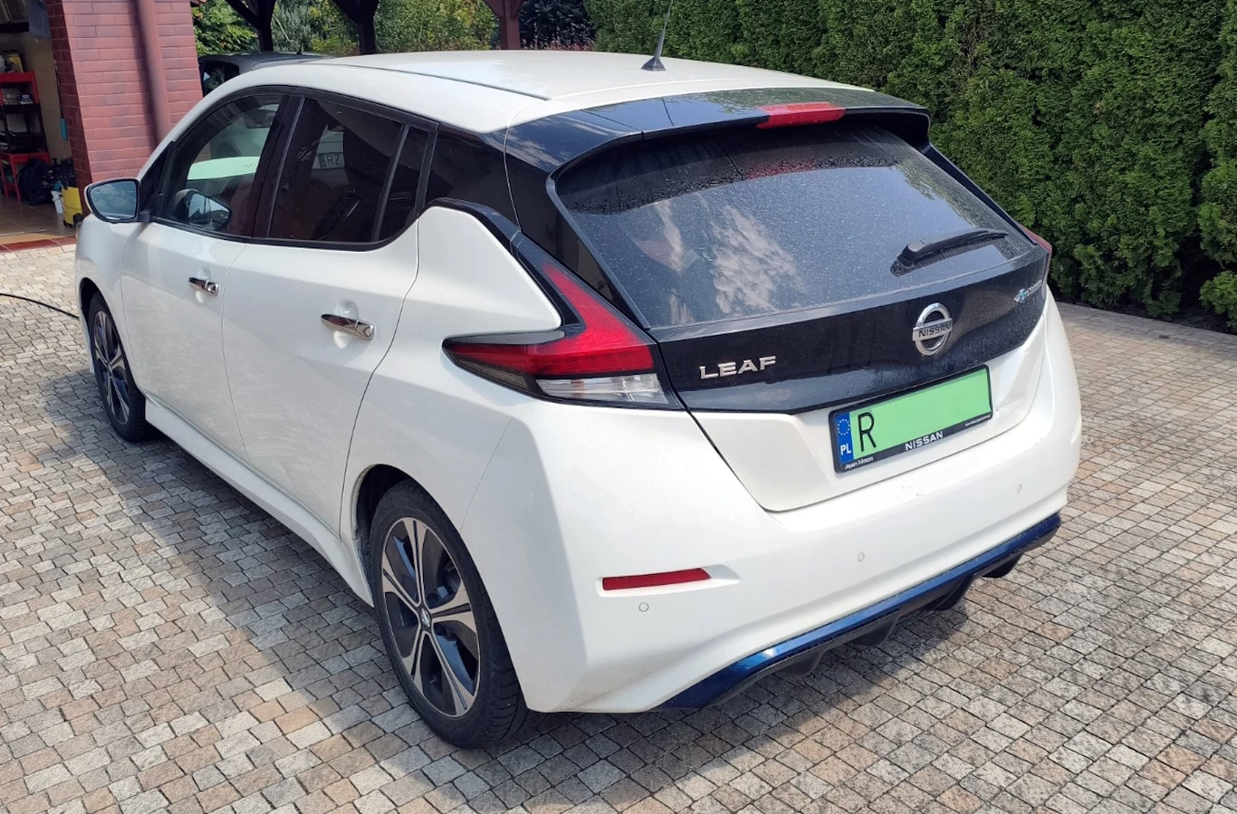Biały Nissan Leaf zaparkowany na brukowanym podjeździe, tył samochodu z widocznymi nowoczesnymi reflektorami i czarnym dachem, w tle żywopłot i fragment budynku.