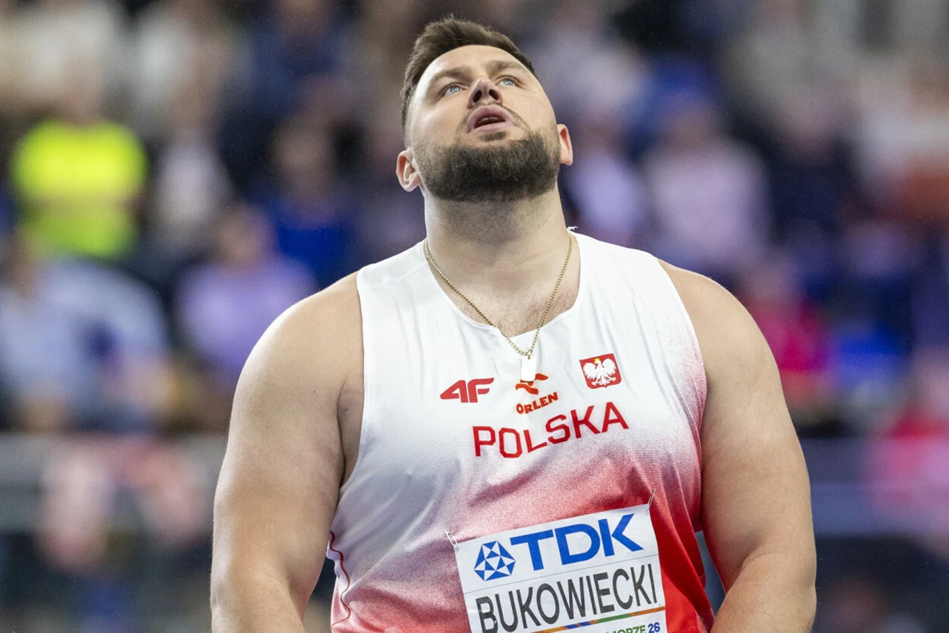 Zawodnik w biało-czerwonym stroju z napisem Polska i identyfikatorem startowym stoi na arenie sportowej, patrząc w górę z wyrazem skupienia na twarzy.