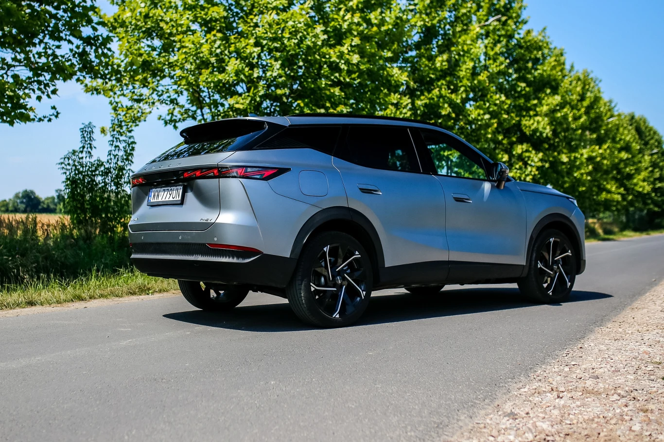 Stylistyka Omody 7 może się podobać. Uwagę zwraca szczególnie nietypowy wzór 20-calowych felg Srebrny SUV o nowoczesnym designie parkuje na asfaltowej drodze w otoczeniu zielonych drzew.
