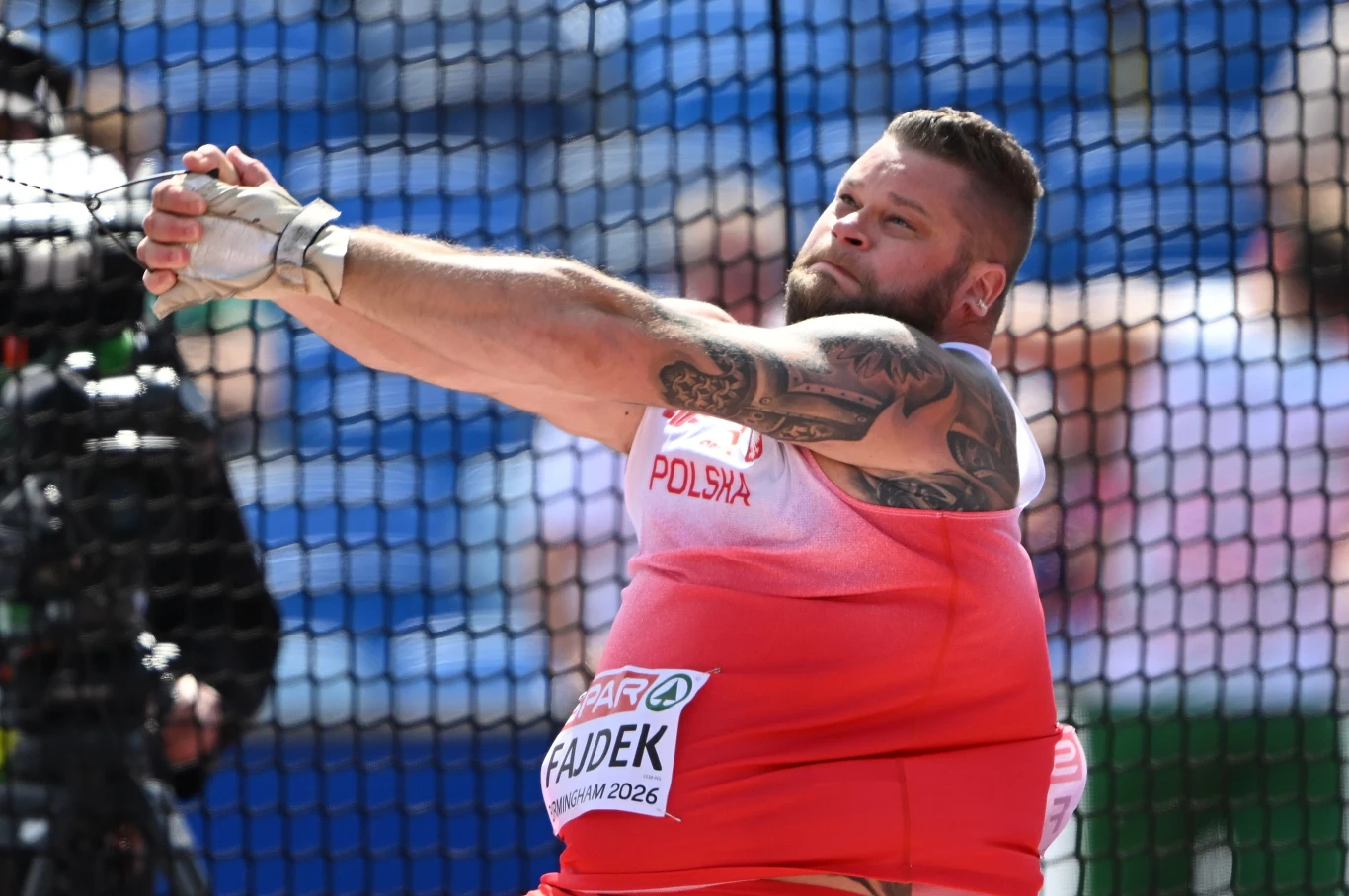 Paweł Fajdek Polski lekkoatleta wykonujący rzut młotem podczas zawodów, skupiony na technice, z widocznymi tatuażami na rękach i ubrany w sportowy strój reprezentacji Polski.