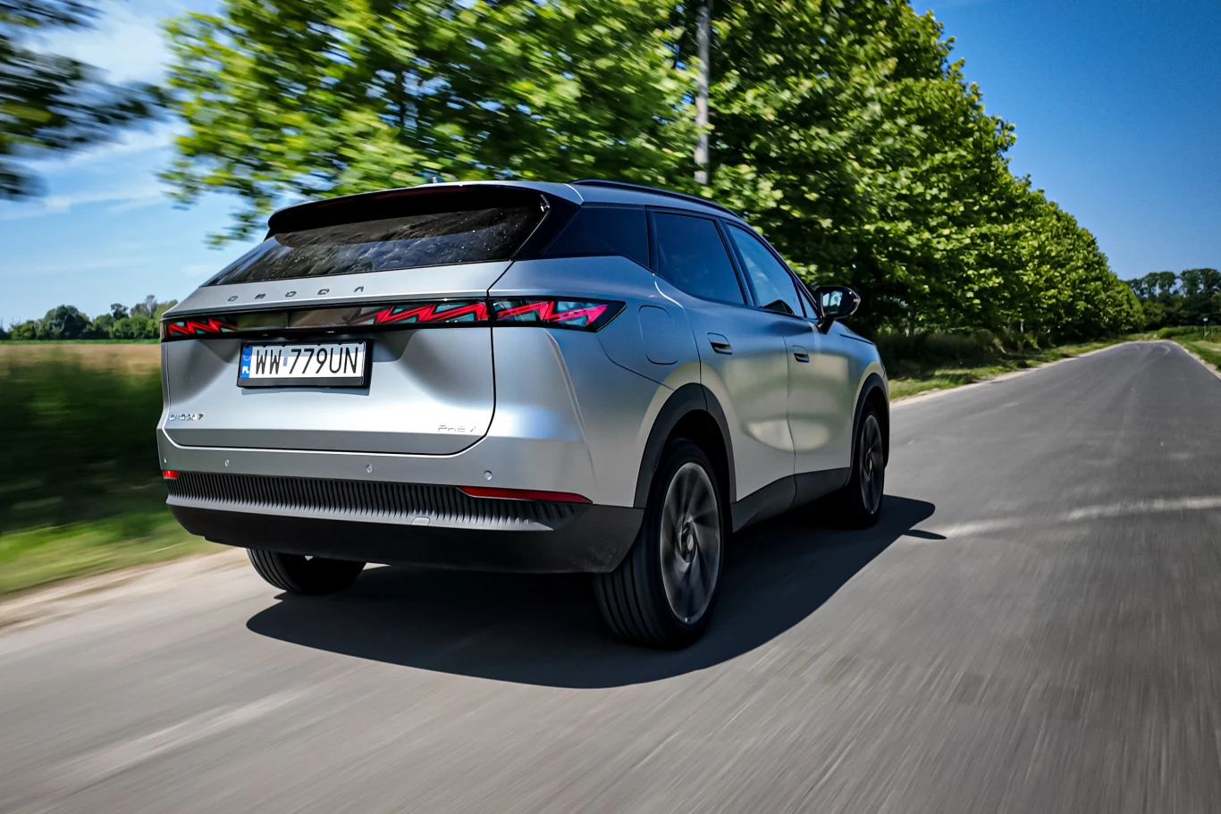 Omoda 7 nieźle się prowadzi, ale zapewnia zbyt niski komfort jazdy jak na dużego SUV-a Srebrny SUV marki Peugeot jedzie asfaltową drogą otoczoną zielonymi drzewami w słoneczny dzień.