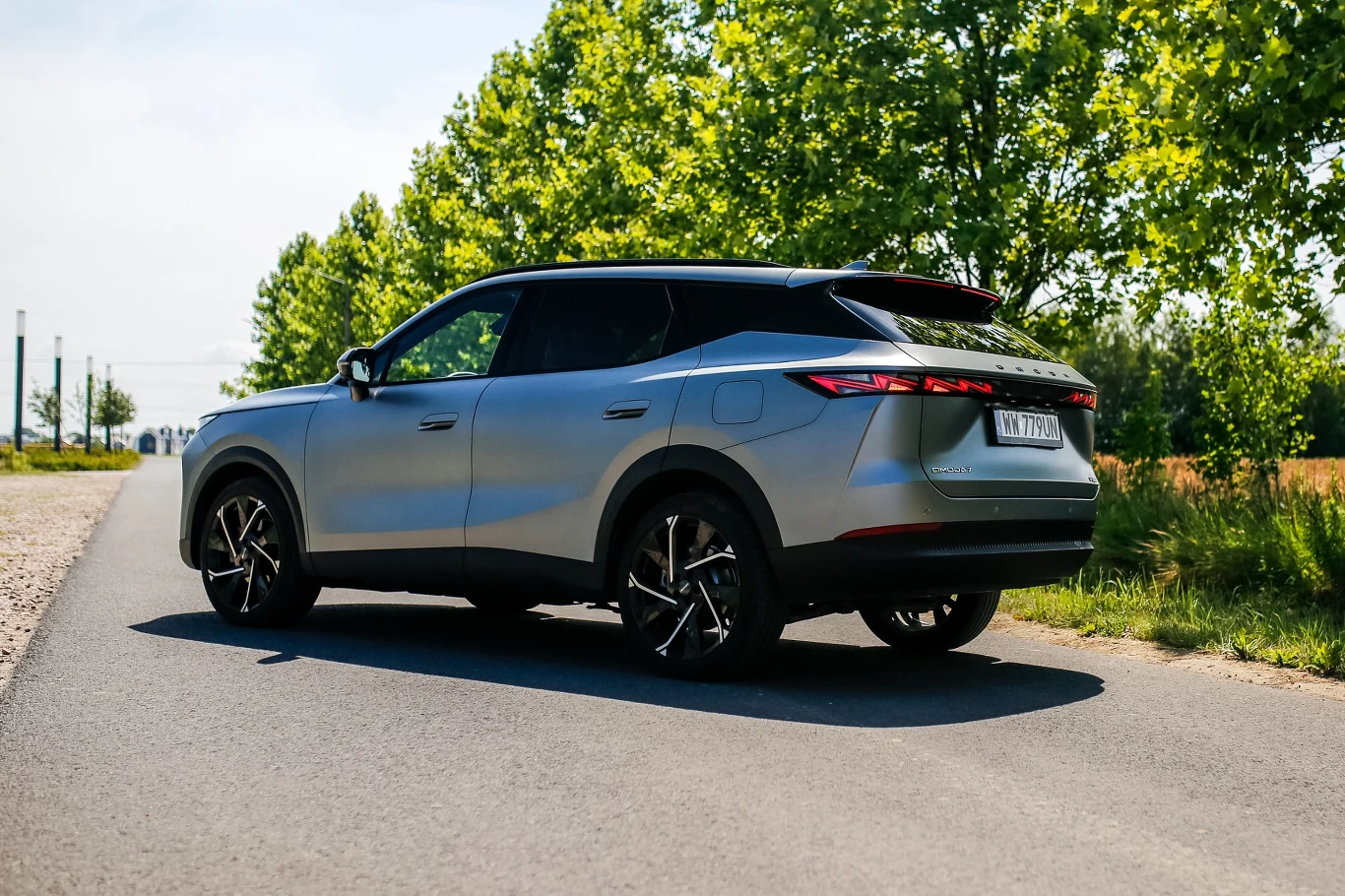 Ceny Omody 7 Super Hybrid zaczynają się od 170 tys. zł Srebrny SUV zaparkowany na asfaltowej drodze, otoczony zielonymi drzewami w słoneczną pogodę.