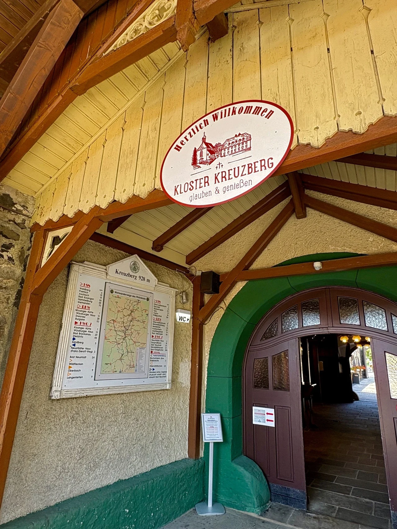Kloster Kreuzberg w Niemczech Wejście do budynku oznaczone czerwonym szyldem z napisem 'Kloster Kreuzberg', pod łukiem drewnianego daszku, obok umieszczona duża mapa oraz plan informacyjny.