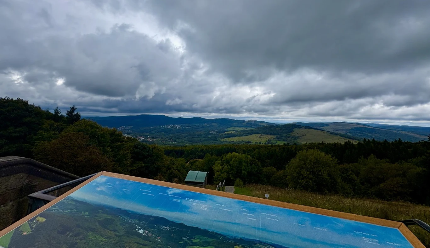 Widoki z góry Kreuzberg robią wrażenie nawet wtedy, gdy pogoda nie dopisuje Rozległa panorama górskich wzgórz pokrytych lasem, z tablicą informacyjną na pierwszym planie i ciemnymi chmurami na niebie.