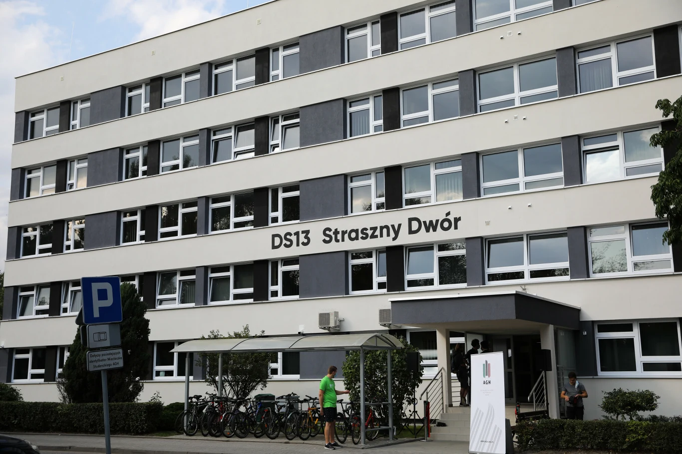Czterokondygnacyjny budynek akademika DS13 Straszny Dwór z dużą liczbą okien, stojakiem na rowery przed wejściem oraz zadaszoną wiatą, obok której znajduje się kilka osób.