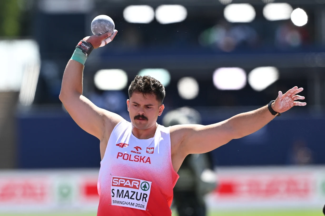 Lekkoatleta w biało-czerwonym stroju z napisem Polska przygotowuje się do pchnięcia kulą, trzymając ją wysoko nad głową, z wyraźnie skupionym wyrazem twarzy.