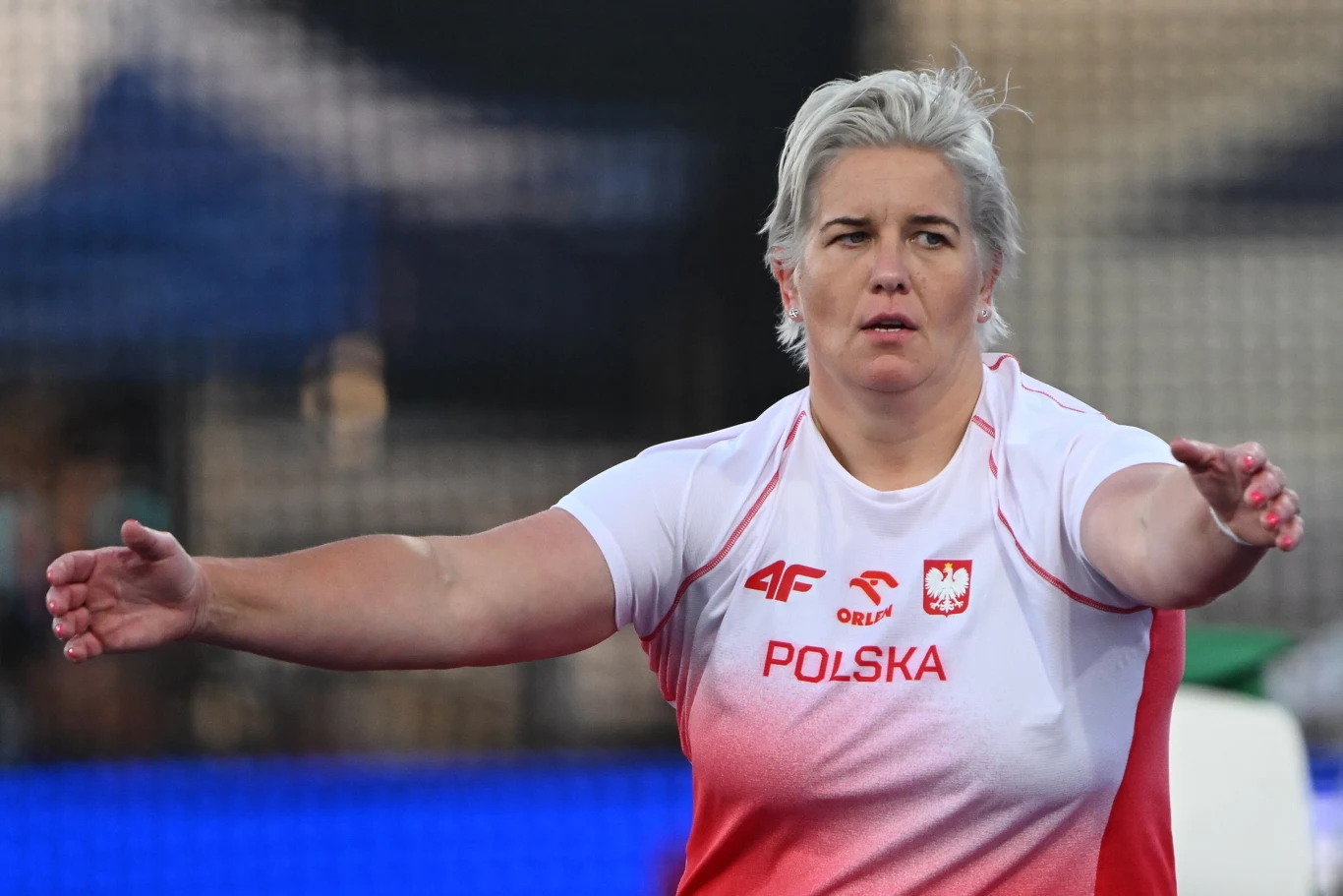 Anita Włodarczyk, Birmingham 2026 Kobieta w sportowej koszulce z napisem 'Polska' i emblematami reprezentacji trzyma rozłożone szeroko ręce, przygotowując się do rzutu na tle stadionu lekkoatletycznego.