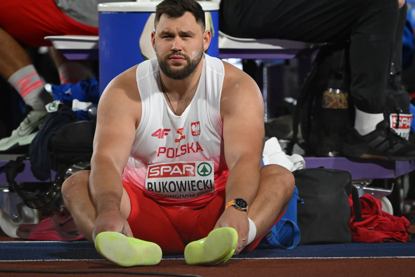 Lekkoatleta w stroju reprezentacji Polski siedzący na podłodze z wyprostowanymi nogami, w tle sprzęt sportowy i torba.