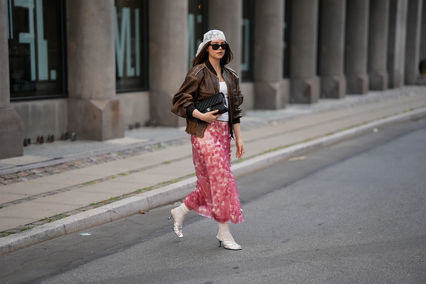 Trendy w modzie na jesień 2026 Kobieta w jasnej czapce z daszkiem i okularach przeciwsłonecznych, ubrana w białą bluzkę, brązową kurtkę i różową spódnicę maxi, idąca chodnikiem z czarną torebką w dłoni.