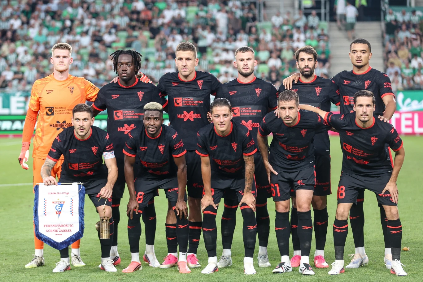 Drużyna Górnika Zabrze w sezonie 2026/2027 Jedenastu piłkarzy ubranych w czarne stroje z czerwonymi akcentami i herbami drużyny pozuje wspólnie na murawie stadionu przed rozpoczęciem meczu piłkarskiego, jeden z zawodników trzyma proporczyk klubowy.