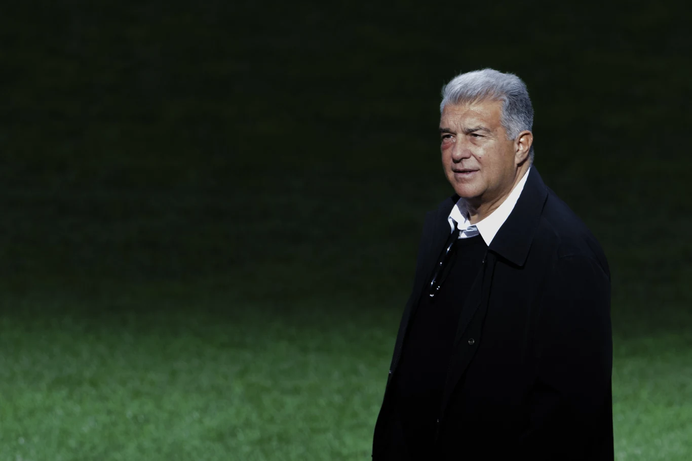 Joan Laporta Mężczyzna w ciemnym płaszczu stoi na tle zielonej murawy, patrząc w stronę aparatu z poważnym wyrazem twarzy.