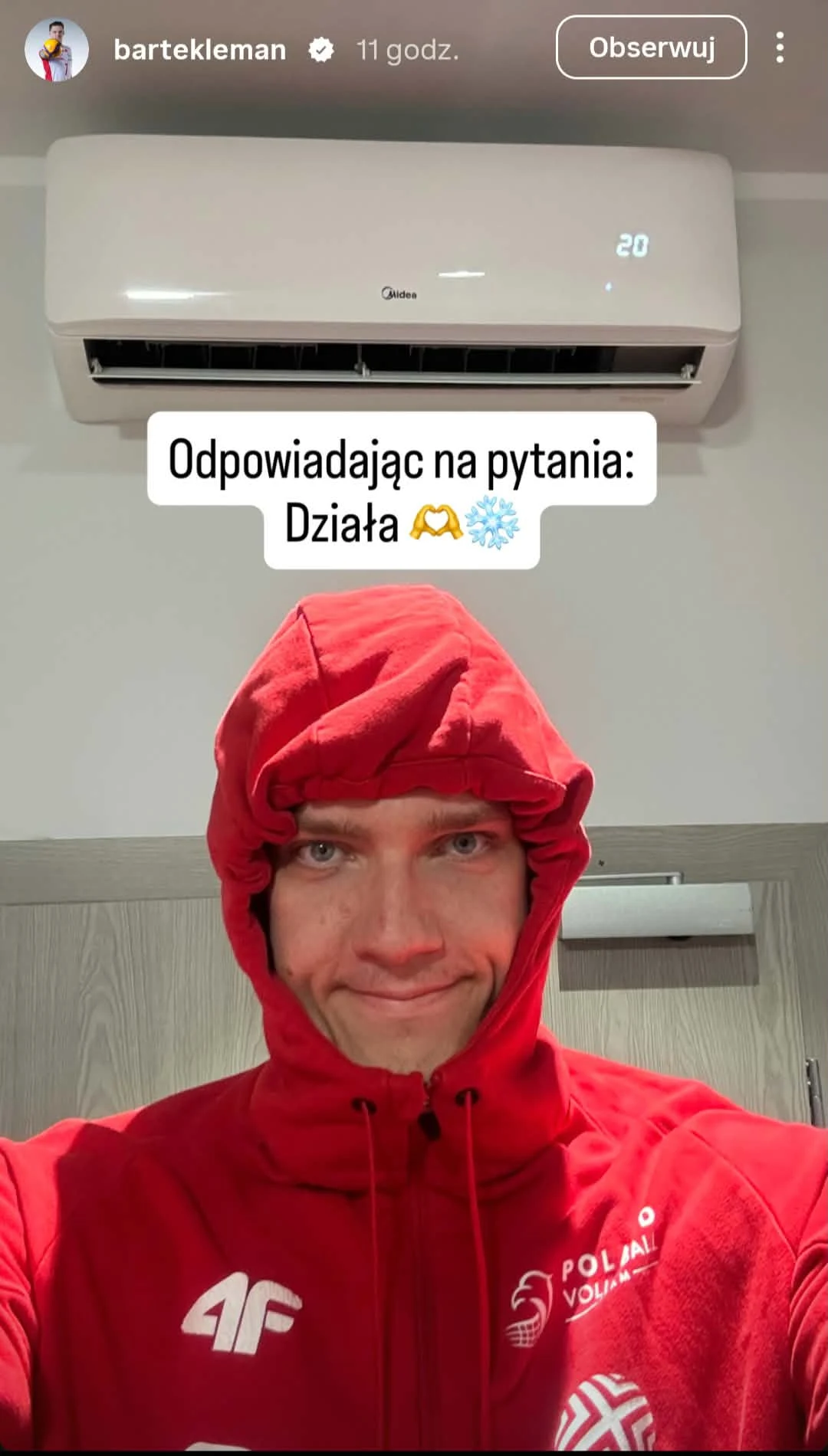 Mężczyzna w czerwonej bluzie z kapturem szczelnie zapiętej pod brodą stoi pod włączonym klimatyzatorem, na którym widnieje temperatura 20 stopni.