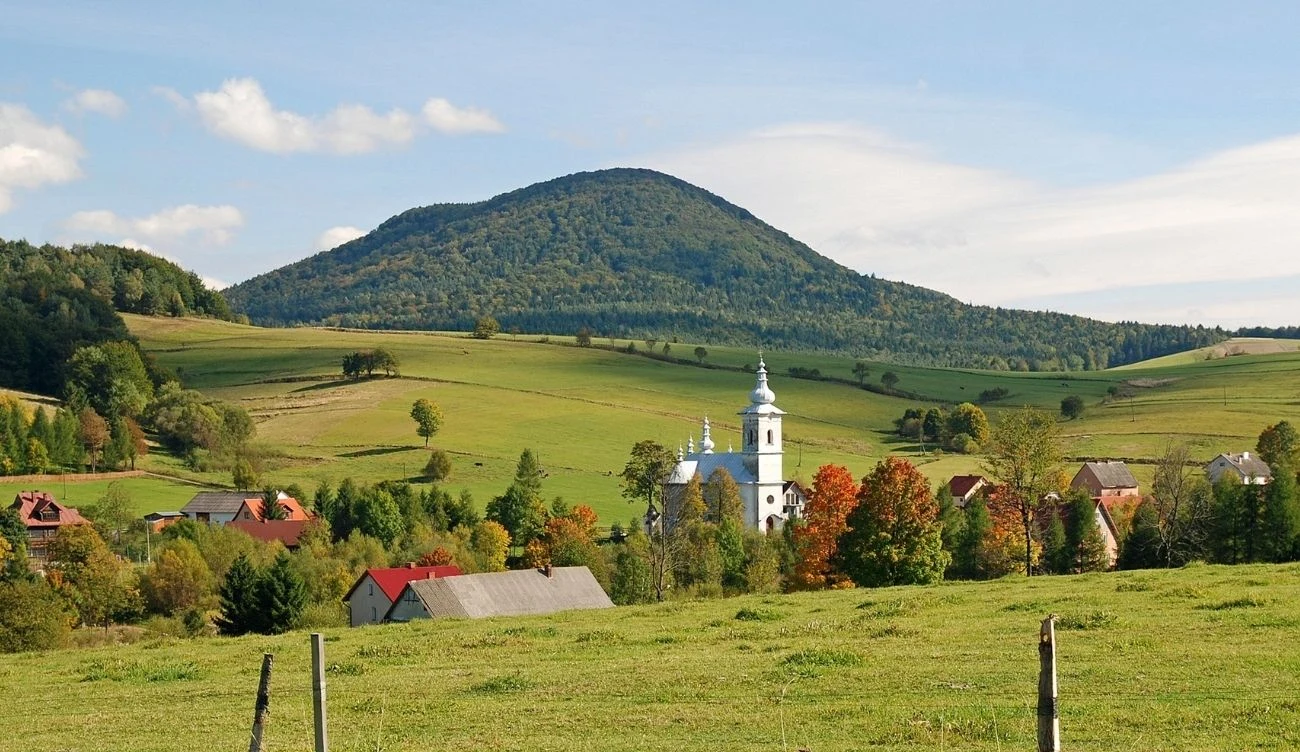 Nowy szlak na Lackową pozwala ominąć strome podejście Wieże kościoła wśród drzew, zabudowania wsi na tle zielonych łąk i zalesionego wzniesienia Lackowej.