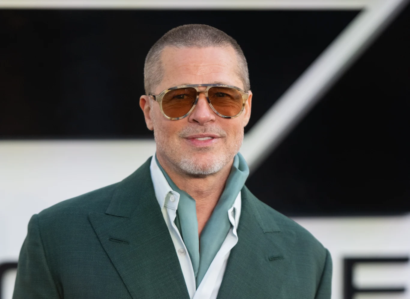 Brad Pitt na premierze "F1" Brad Pitt w zielonej marynarce, jasnoniebieskiej koszuli i okularach, na tle geometrycznych kształtów.