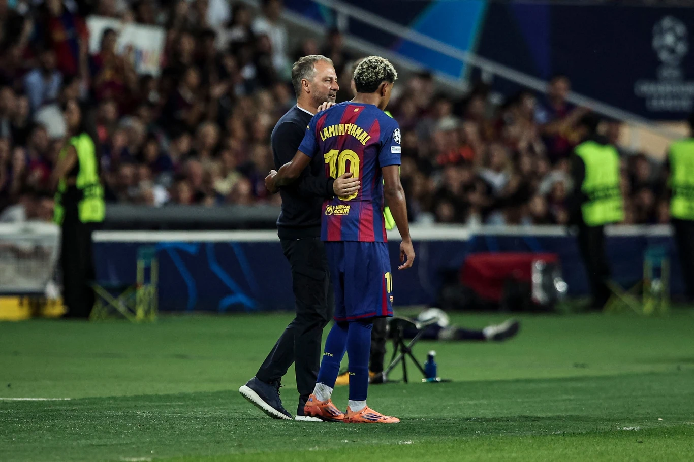 Trener rozmawia z piłkarzem drużyny FC Barcelona ubranym w koszulkę z numerem 10 podczas meczu na murawie stadionu, w tle widoczni ochroniarze i kibice.
