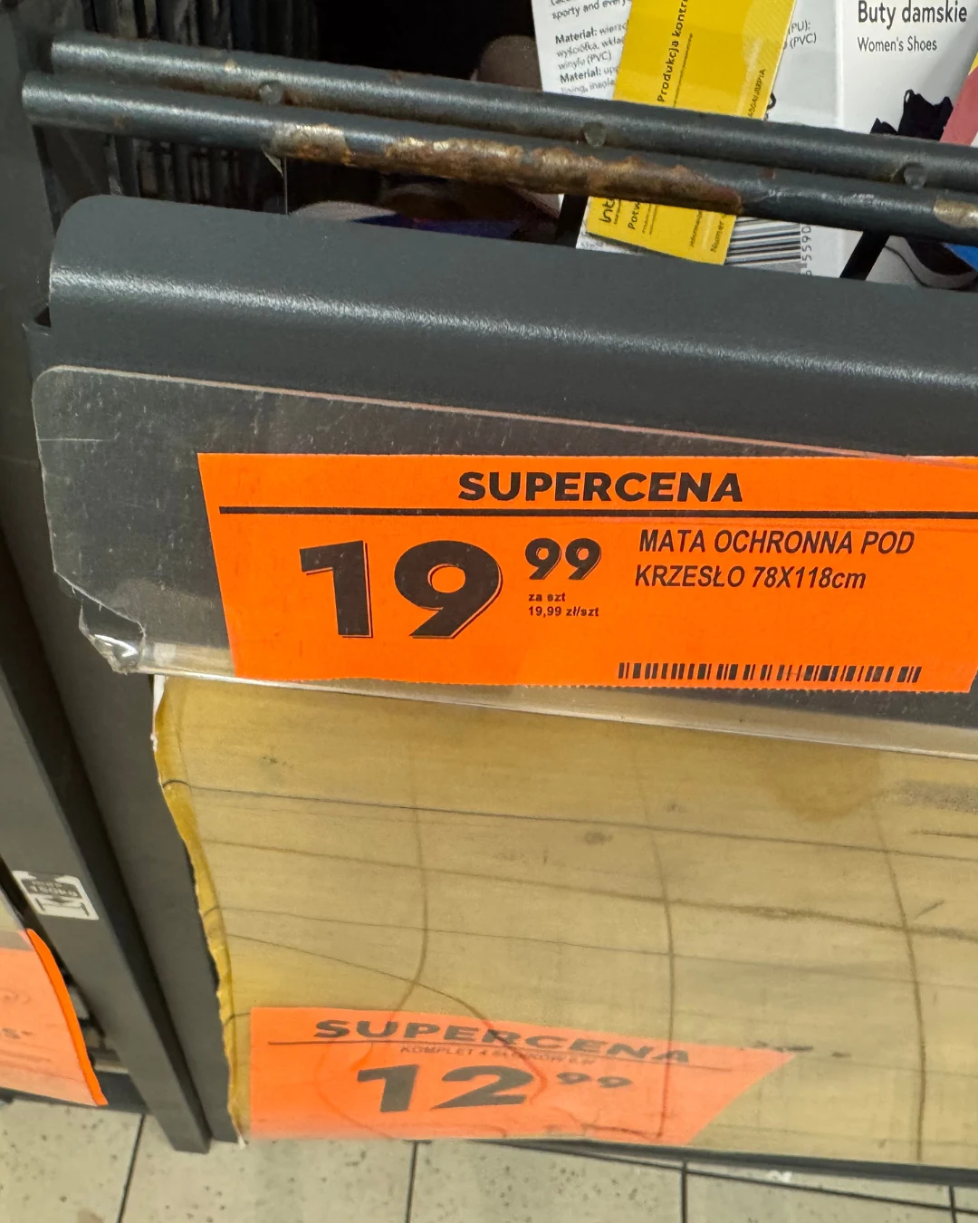 Cena maty ochronnej Pomarańczowa etykieta z ceną 19,99 zł oznaczająca matę ochronną pod krzesło o wymiarach 78x118 cm, umieszczona na półce sklepowej nad transparentną matą.