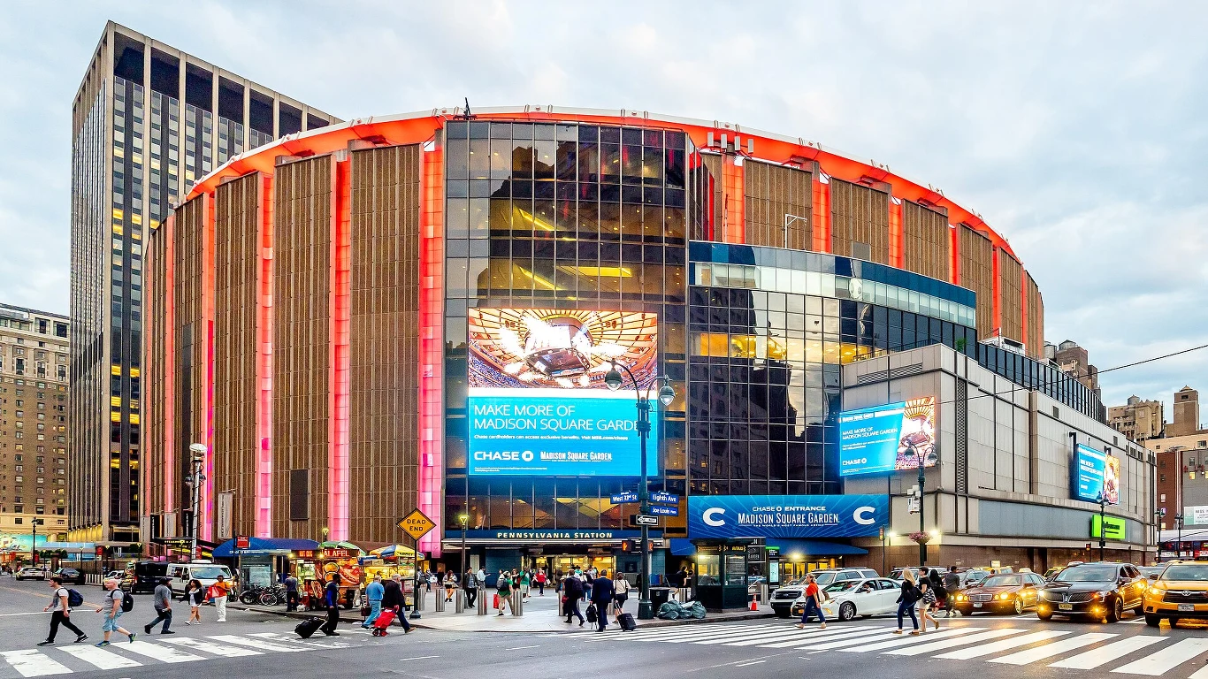 Okrągły budynek Madison Square Garden z podświetlonymi panelami elewacyjnymi i dużymi ekranami LED wyświetlającymi reklamy, otoczony przez ruchliwe ulice i grupy pieszych.