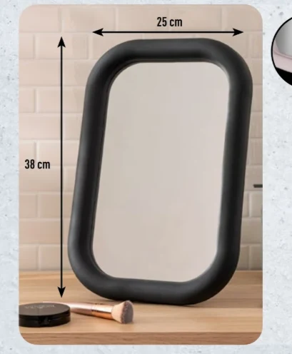 Nowoczesne lustro za 22,99 zł Owalne lustro o czarnej, matowej ramie o wymiarach 38 na 25 cm ustawione na drewnianym blacie, obok którego leży pędzel do makijażu i pudełko z kosmetykiem.