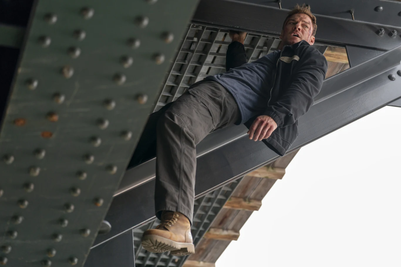 Alan Ritchson w serialu "Reacher" Mężczyzna w kurtce i jasnych butach zwisa z jednej ręki pod stalową konstrukcją mostu, patrząc w stronę kamery; jego wyraz twarzy sugeruje napięcie lub wysiłek.