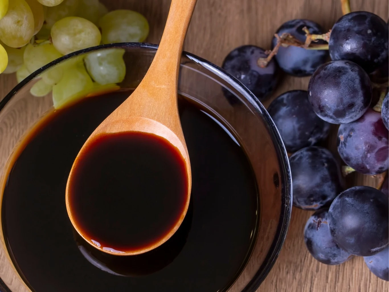 Ocet winogronowy rozcieńczony z wodą ma działanie wspierające trawienie oraz pomaga dbać o prawidłowy poziom cukru we krwi Drewniana łyżka z ciemnym octem winogronowym w miseczce, obok świeże zielone i ciemnofioletowe winogrona.