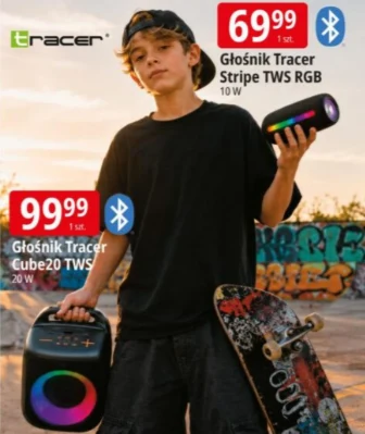 Głośniki z RGB od 69,99 zł Chłopak w czarnej koszulce i czapce z daszkiem trzyma w jednej ręce kolorowy głośnik bezprzewodowy, a w drugiej większy głośnik z podświetleniem RGB i deskorolkę; w tle można dostrzec graffiti na murze.