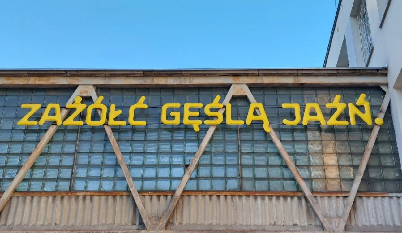 To zdanie zawiera w sobie wszystkie polskie znaki diakrytyczne Żółty napis „ZAŻÓŁĆ GĘŚLĄ JAŹŃ” na tle przeszklonych okien industrialnego budynku z metalową konstrukcją.